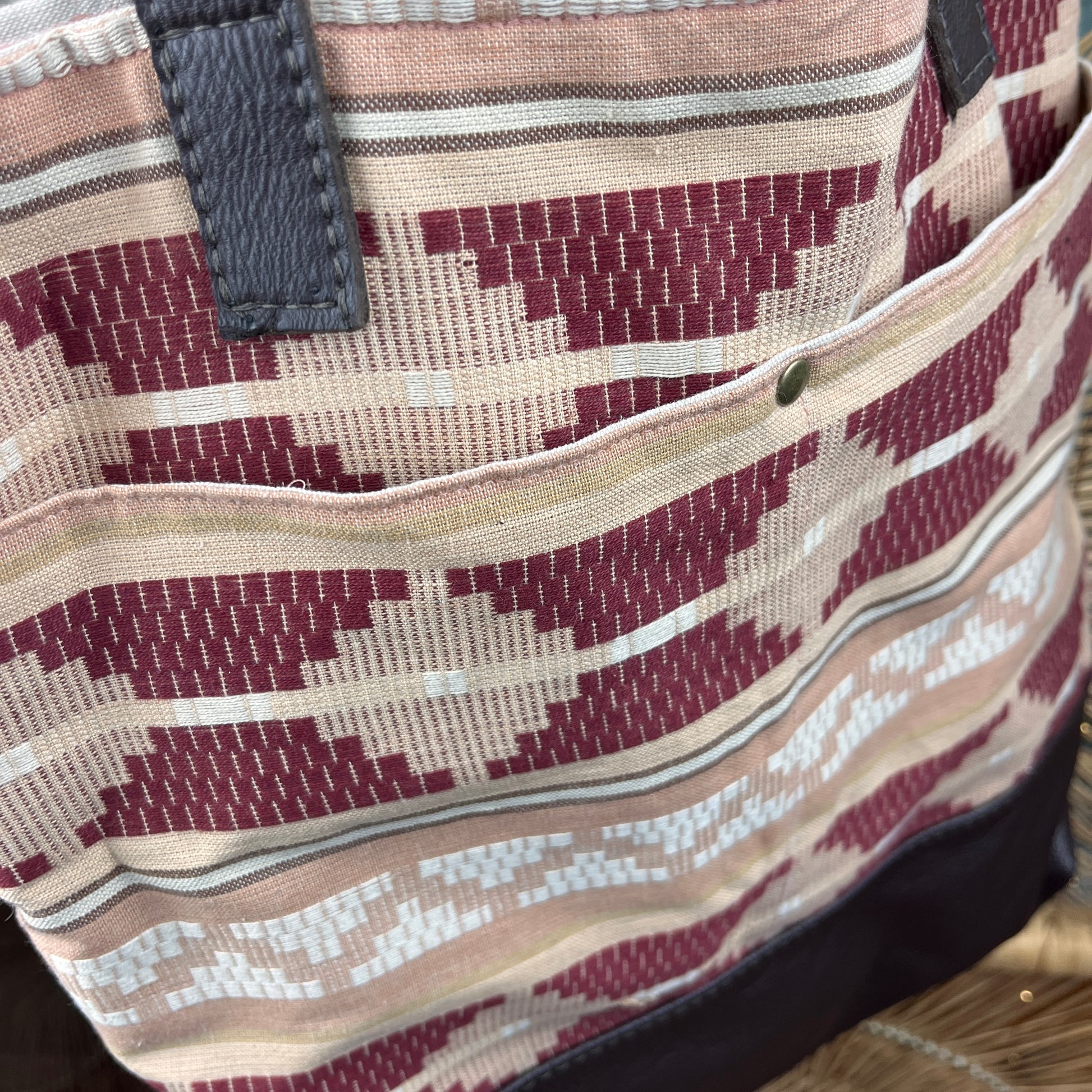 Close-up of a woven tote bag with a geometric ikat pattern in burgundy, beige, and white, featuring a front pocket and dark gray strap attachments.