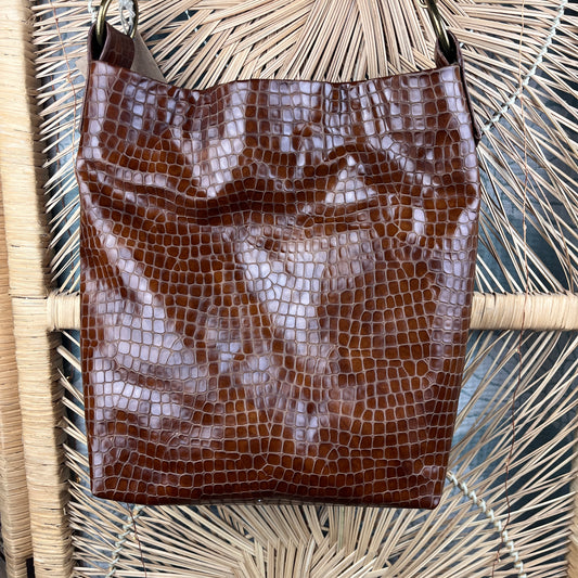 A brown crocodile-embossed leather shoulder bag hanging against a woven straw chair back.