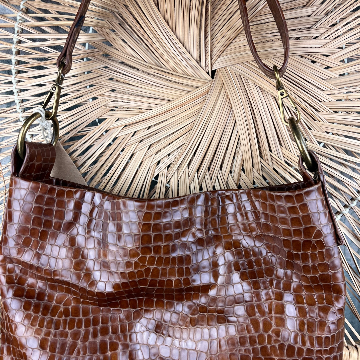 A brown crocodile-embossed leather shoulder bag with gold-tone hardware, shown against a background of dried palm leaves.