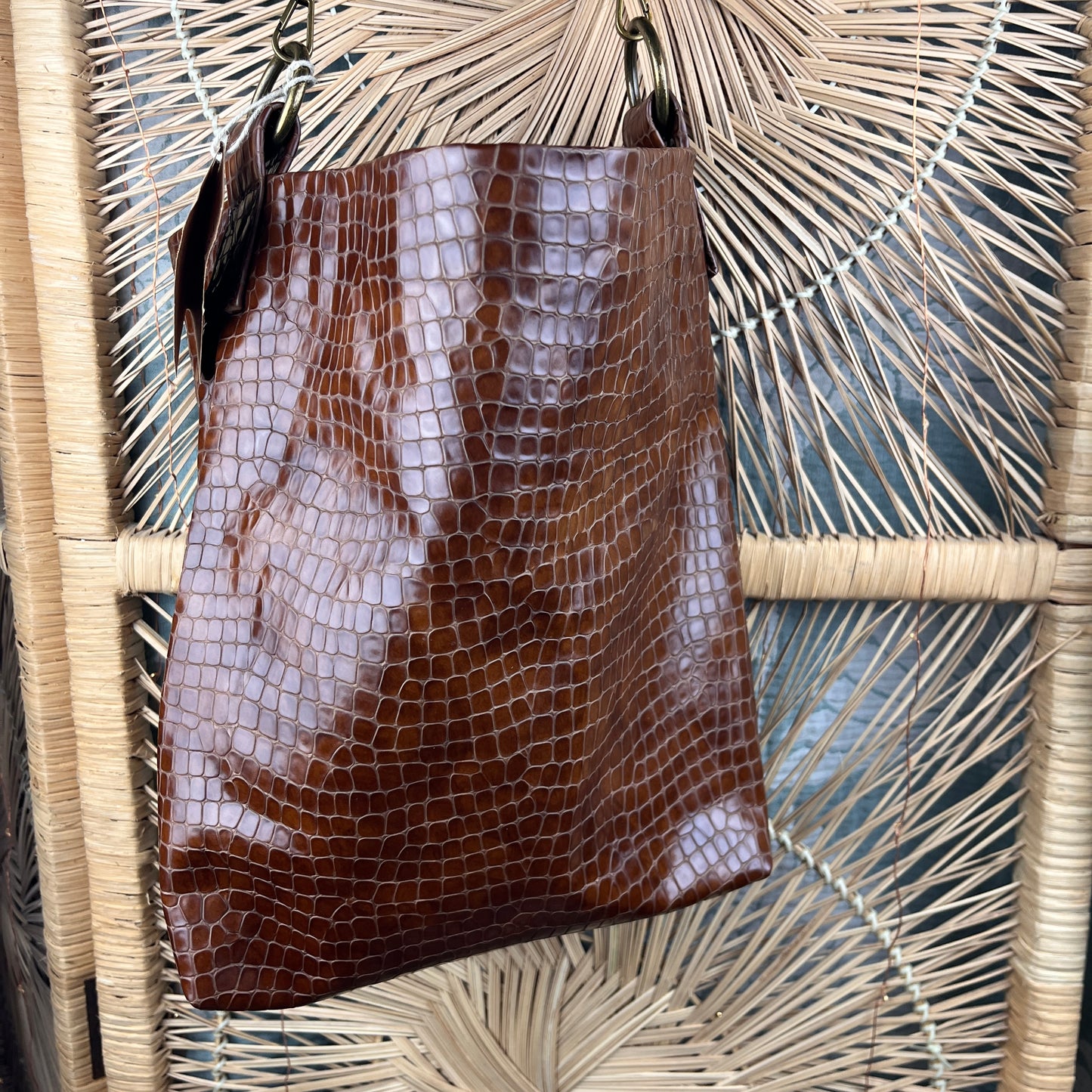 A brown crocodile-embossed leather tote bag hanging against a woven rattan chair back.