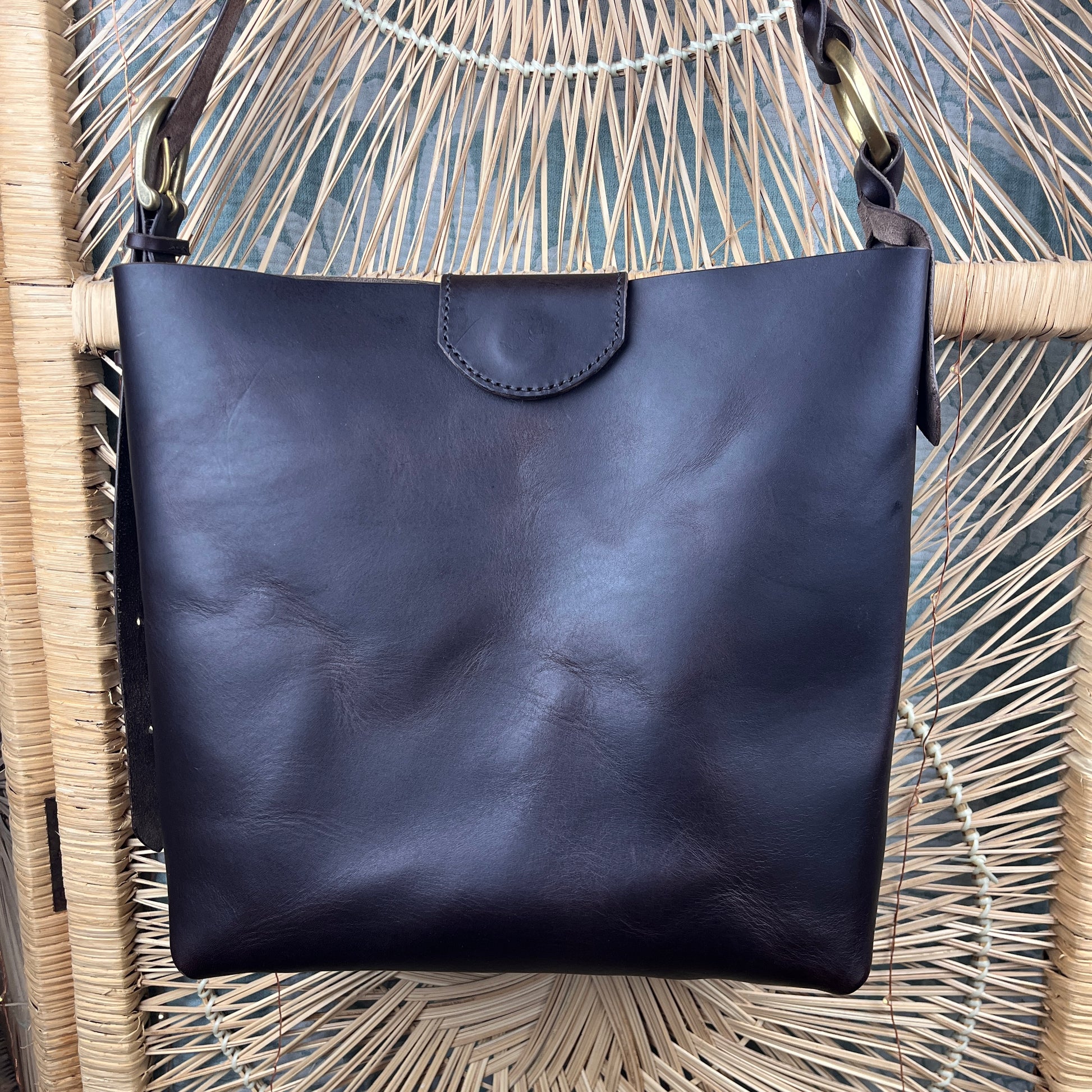 An espresso leather shoulder bag with a flap closure, hanging from a woven rattan chair.
