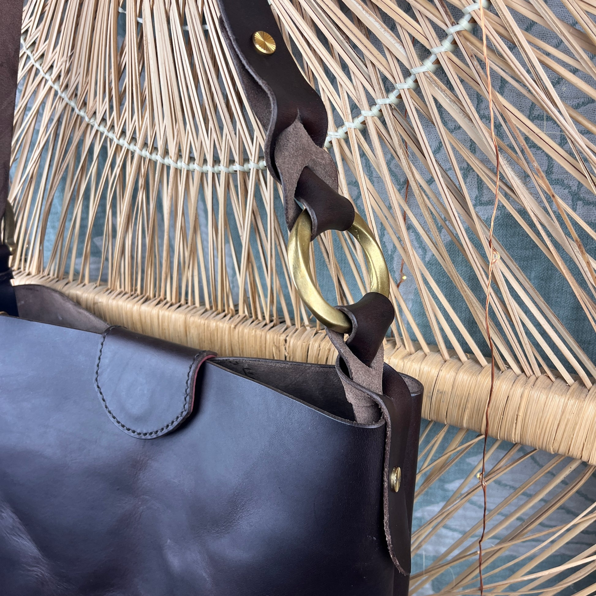 Close-up of a dark brown leather bag with a wide strap featuring a large gold-tone ring and a gray suede accent, set against a woven straw background.