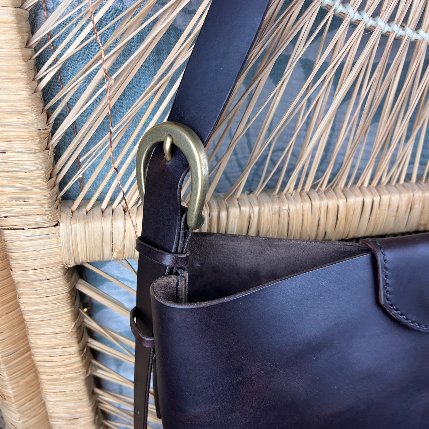Close-up of a dark brown leather shoulder bag with a wide strap and a large antique bronze metal ring, resting against a woven straw chair.