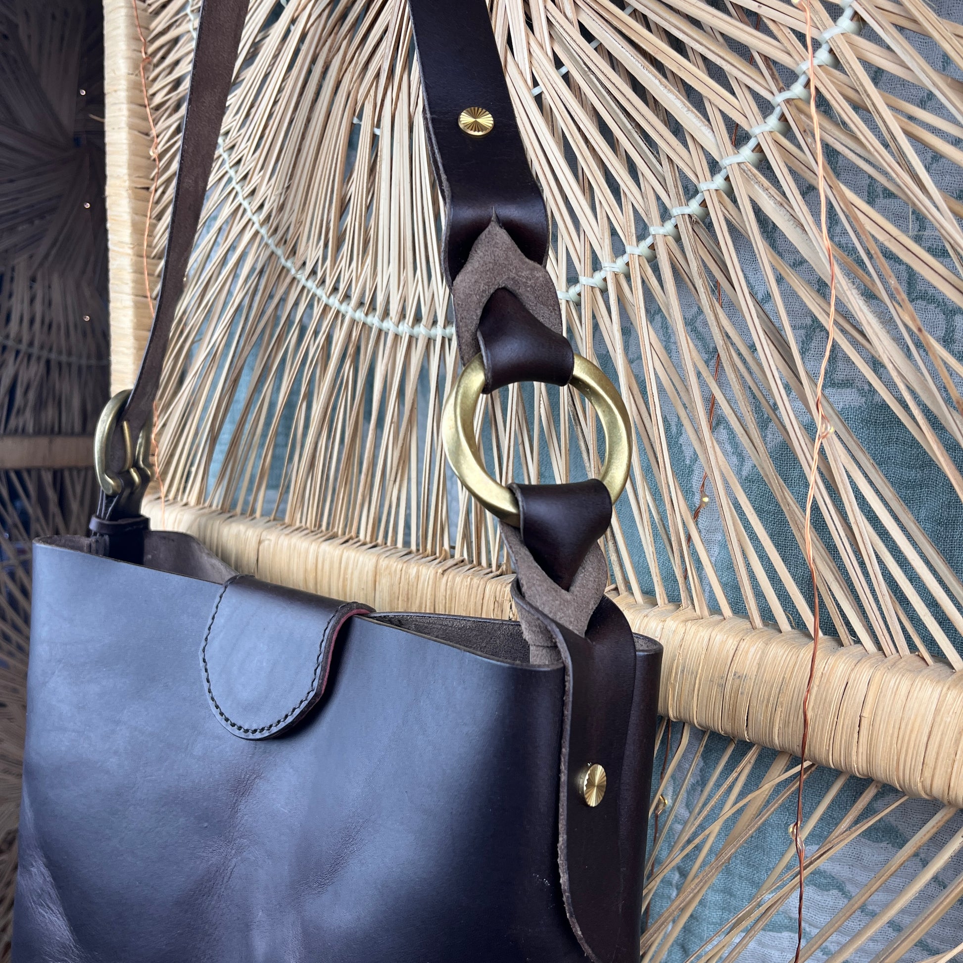 A close-up of a dark brown leather shoulder bag with a wide braided strap and a large brass ring, set against a woven straw background.