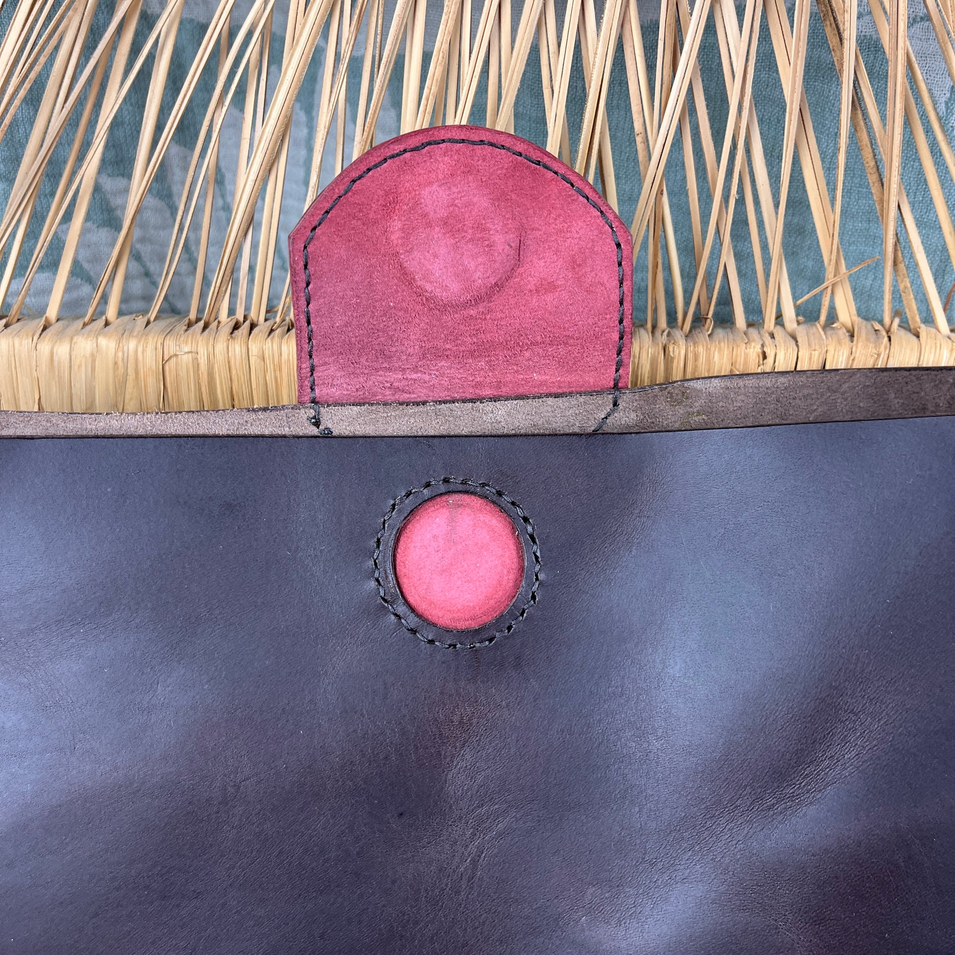 Close-up of a handbag or tote bag showing a dark brown leather lower section, a natural woven straw upper section, and a red leather tab and circular snap closure.