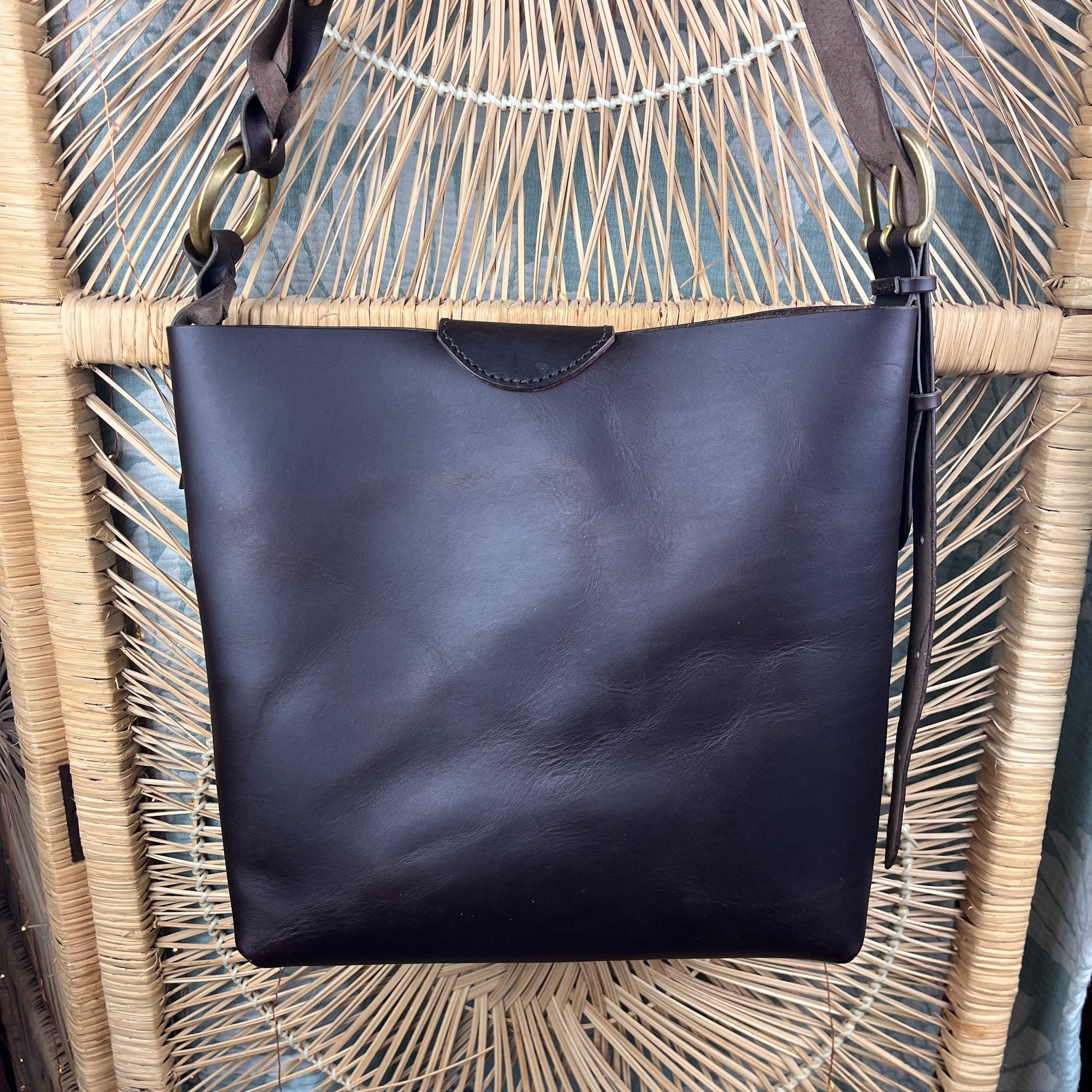 A dark brown leather shoulder bag with a wide top opening, hanging from a woven rattan chair.