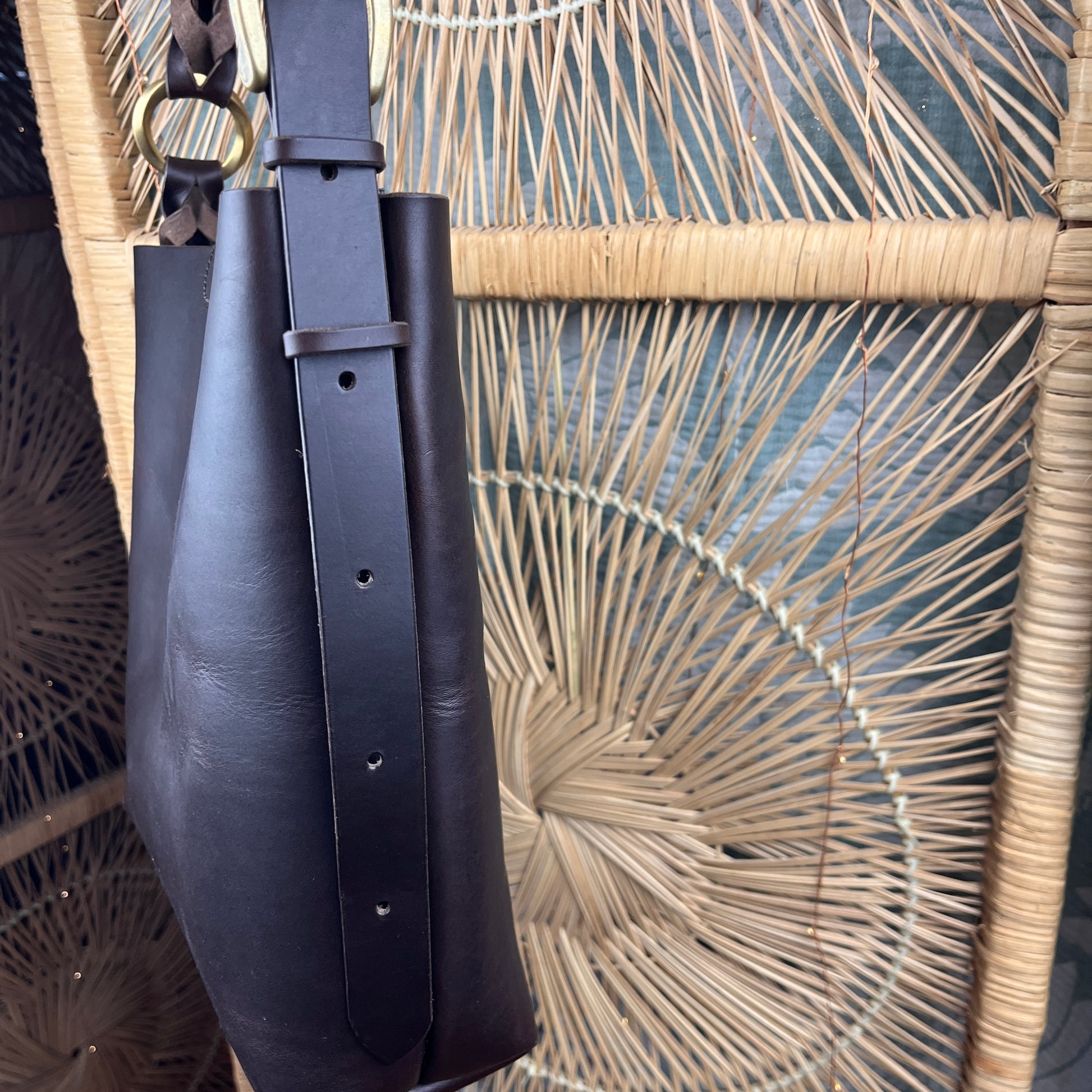 A dark brown leather bucket bag with a wide strap and metal ring hardware, shown hanging in front of a woven rattan screen.
