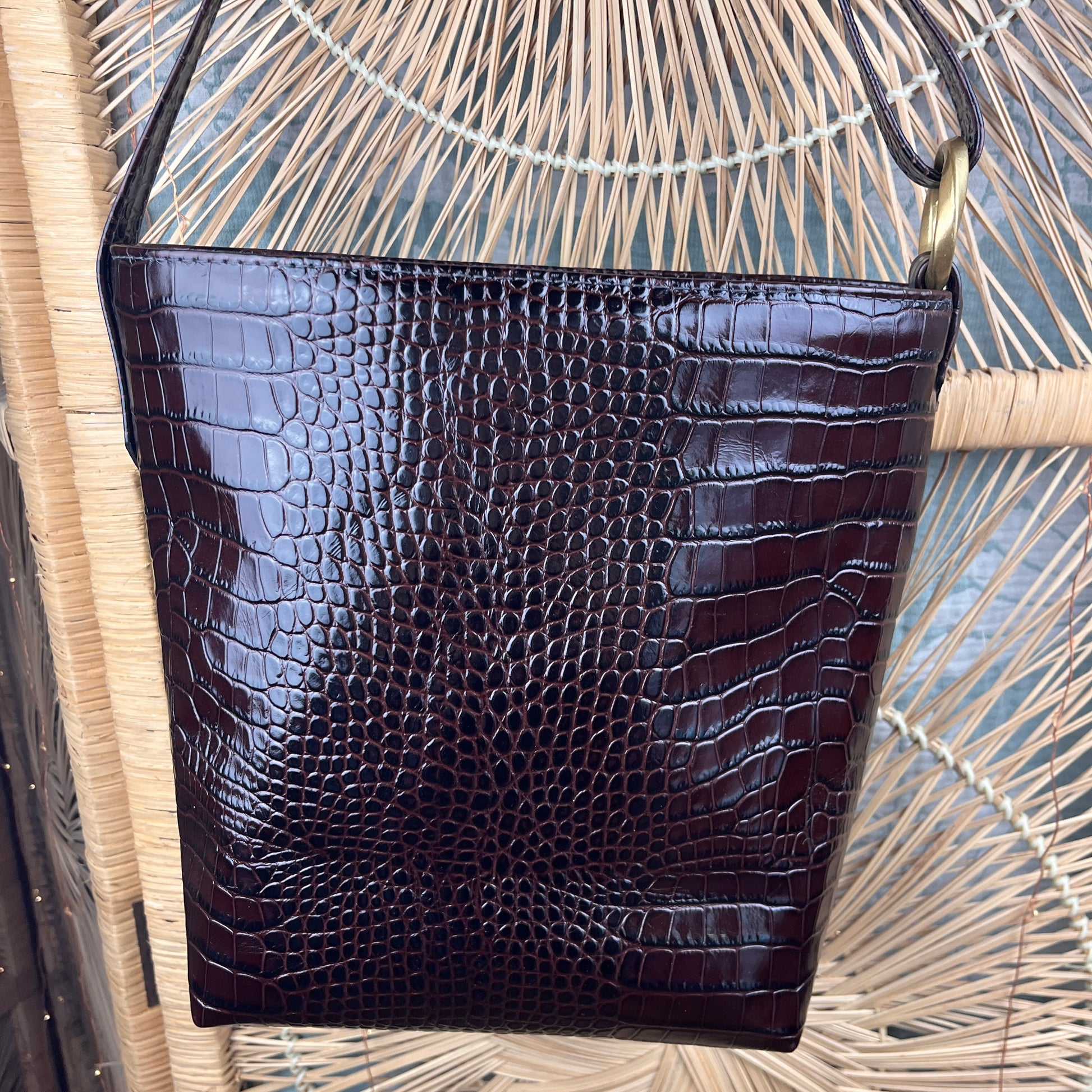 A dark brown crocodile-embossed leather shoulder bag with a gold-tone metal ring, hanging against a woven straw background.