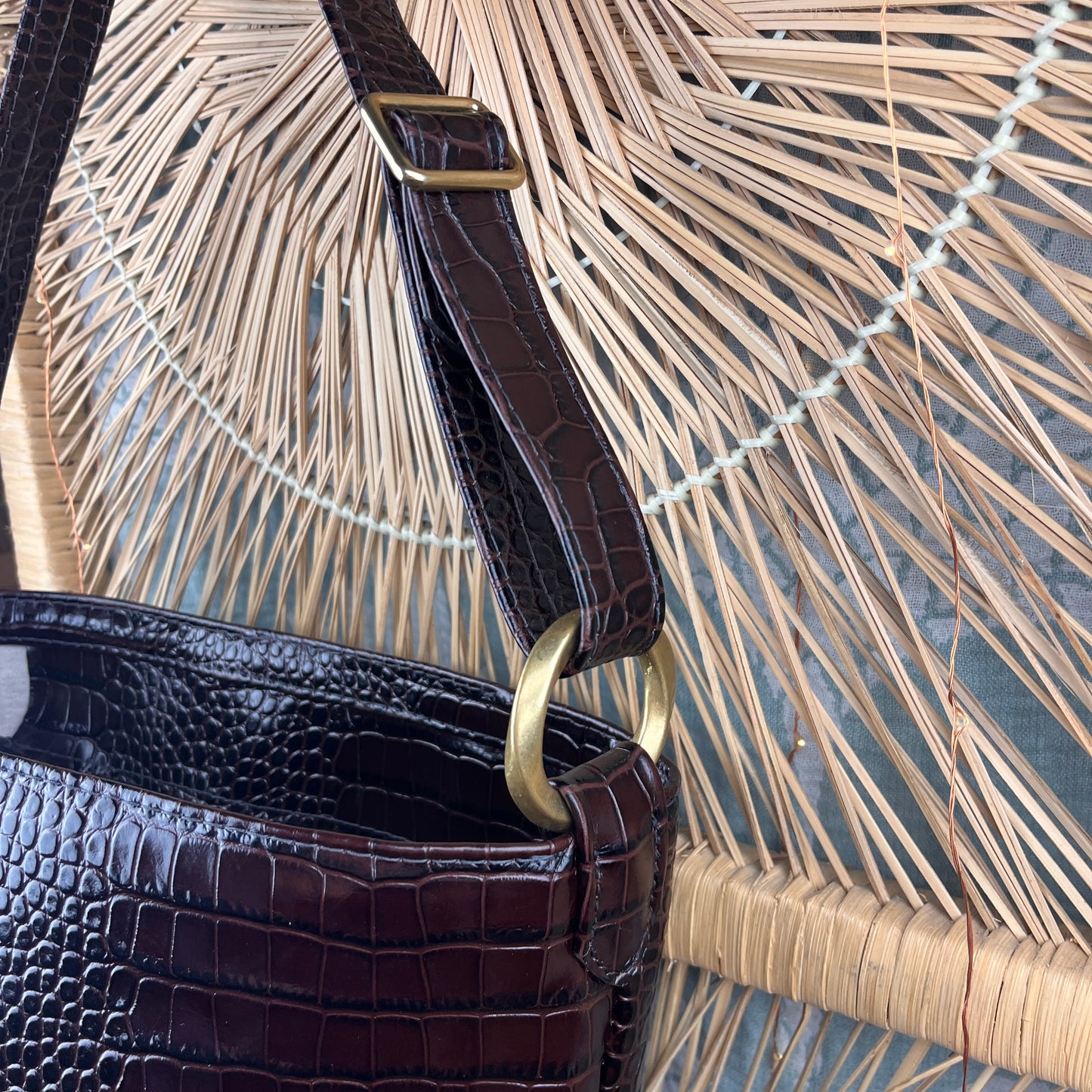 A close-up of a dark brown crocodile-leather handbag with a gold-tone metal ring and strap buckle, set against a woven straw rattan background.