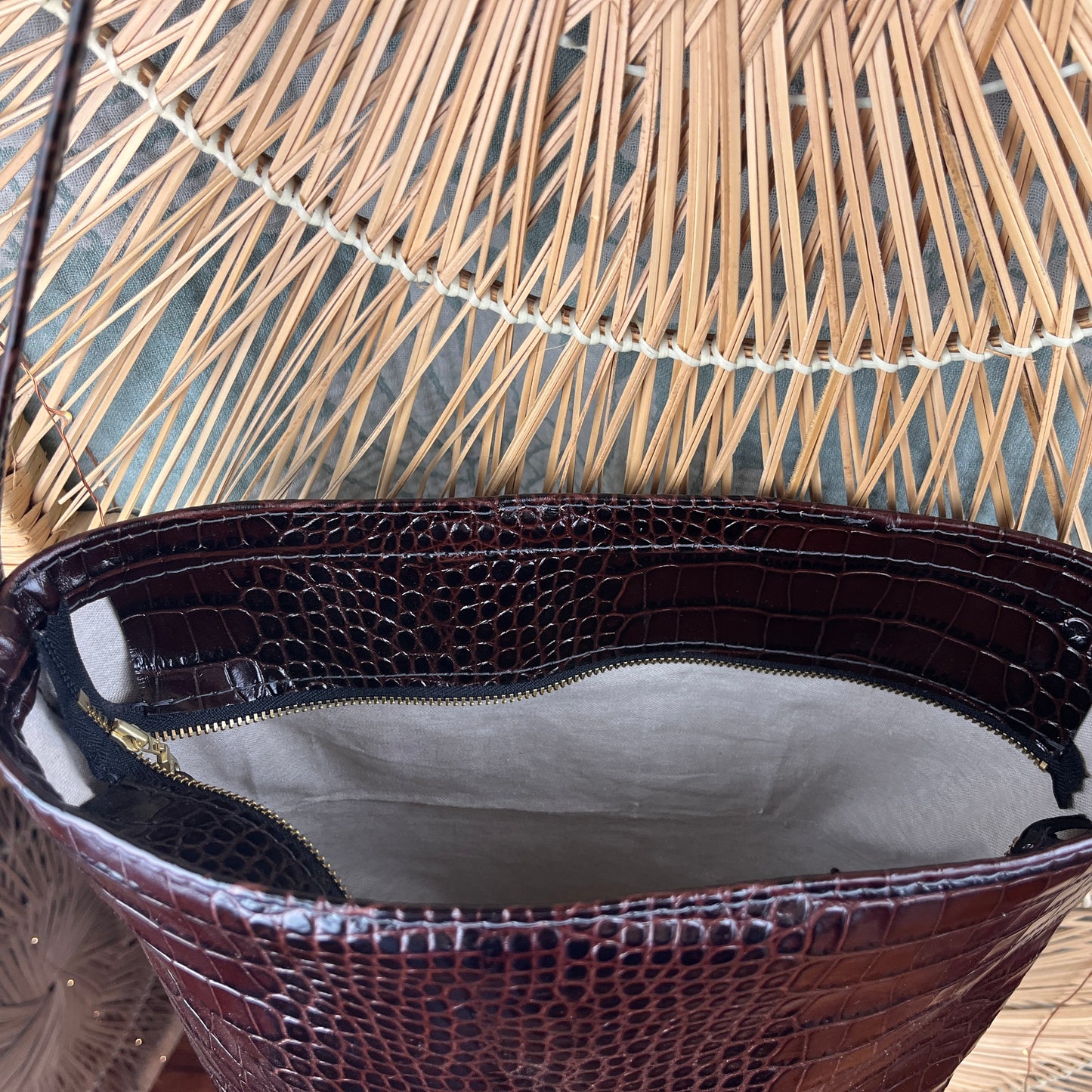 Close-up of the open top of a dark brown crocodile-embossed leather bag, showing a light gray interior lining and zipper, set against a woven straw background.