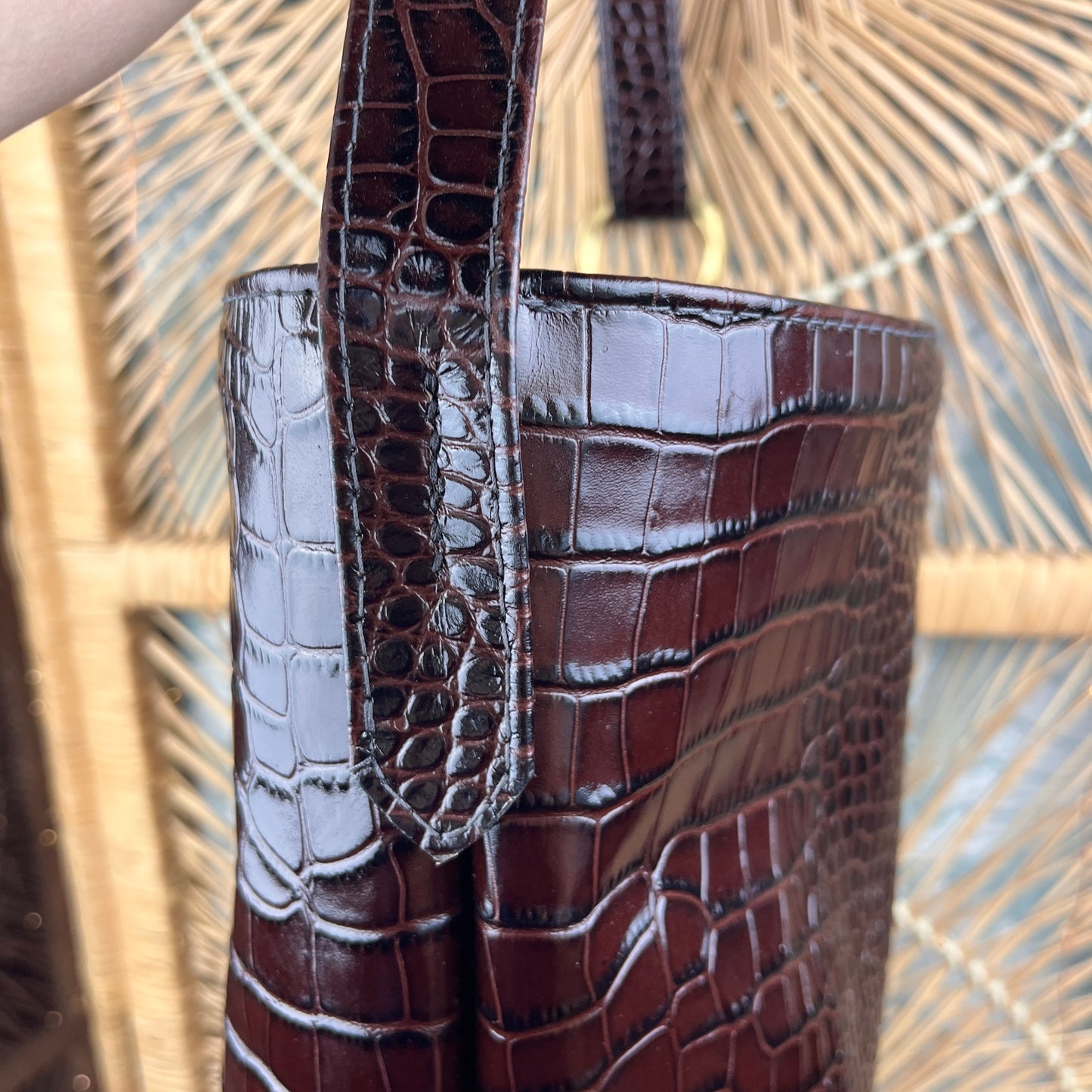 Close-up of a dark brown crocodile-embossed leather bag with a matching strap and gold-tone hardware, set against a woven straw background.