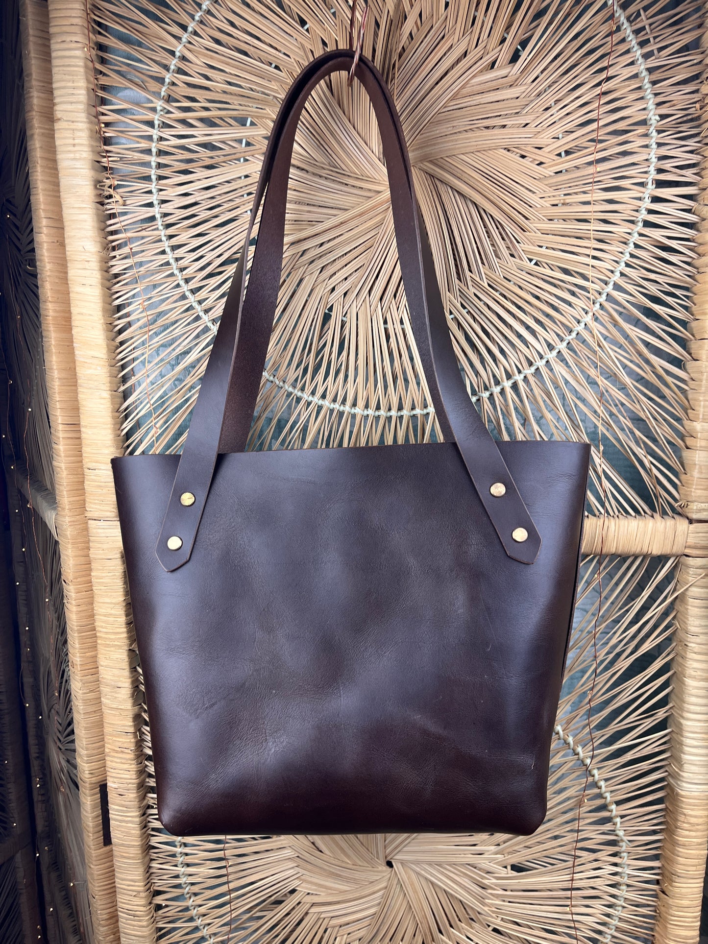 A dark brown leather tote bag with double shoulder straps and brass grommets, hanging against a woven rattan screen.