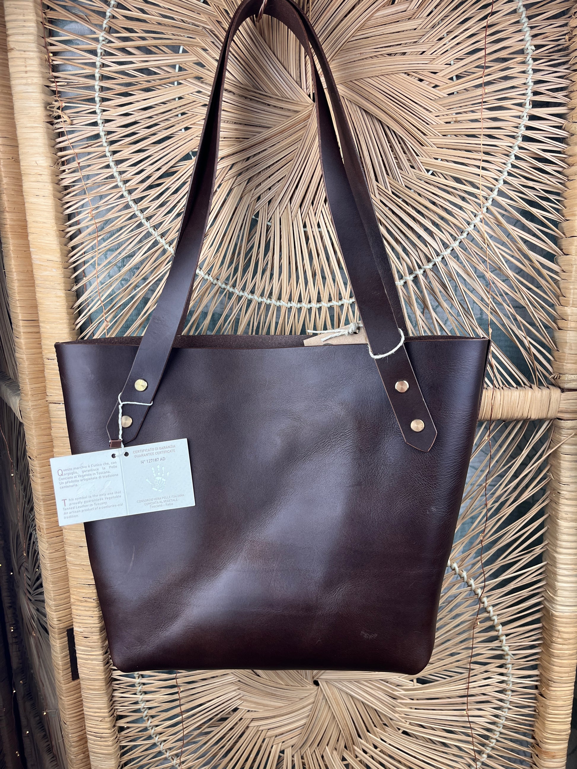 A dark brown leather tote bag with long shoulder straps and brass-tone rivets, hanging in front of a woven rattan screen. A white paper tag is attached to one strap.