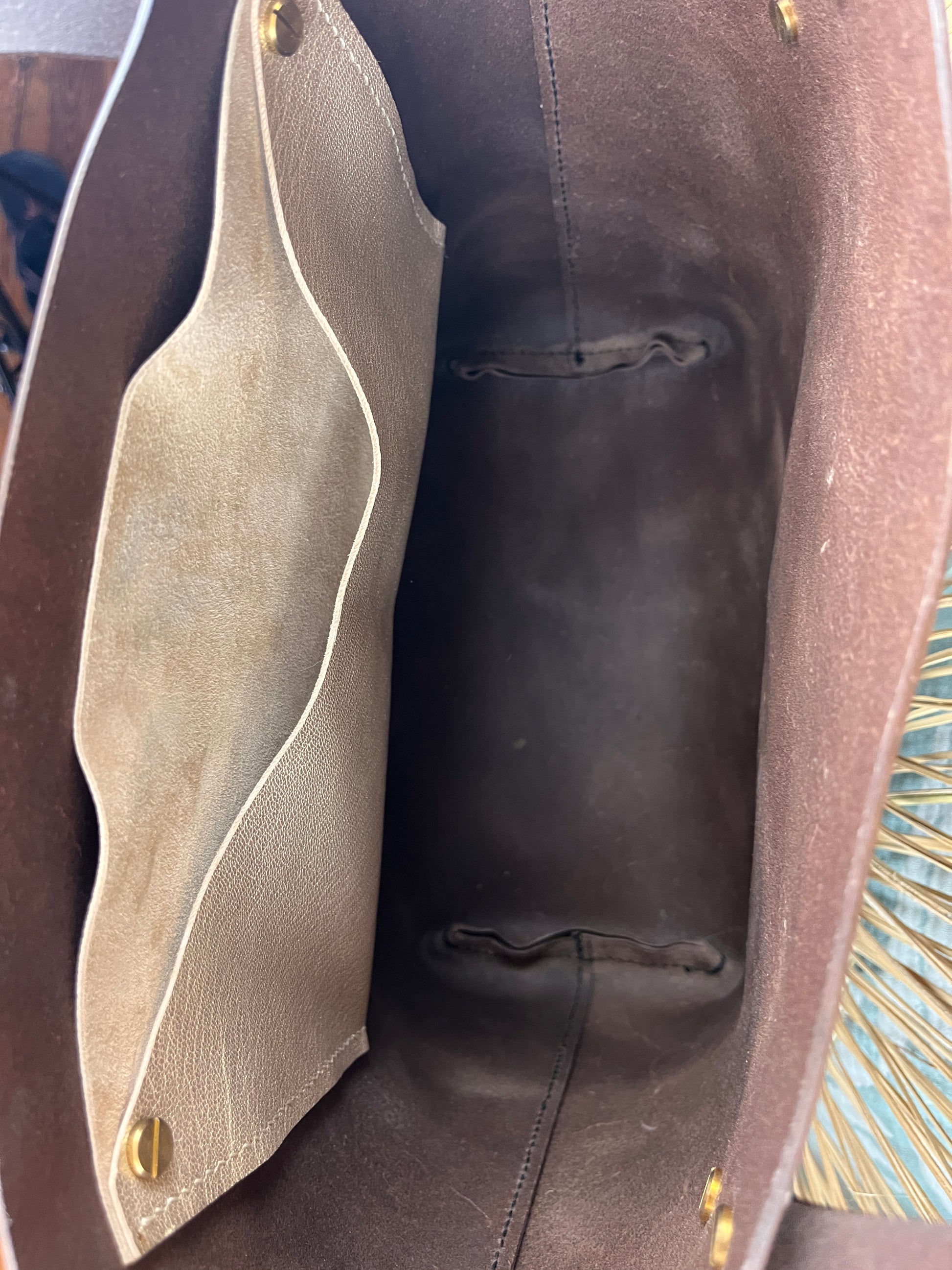 Close-up view inside a brown leather bag, showing a beige suede or nubuck interior organizer panel with gold-tone snap hardware.