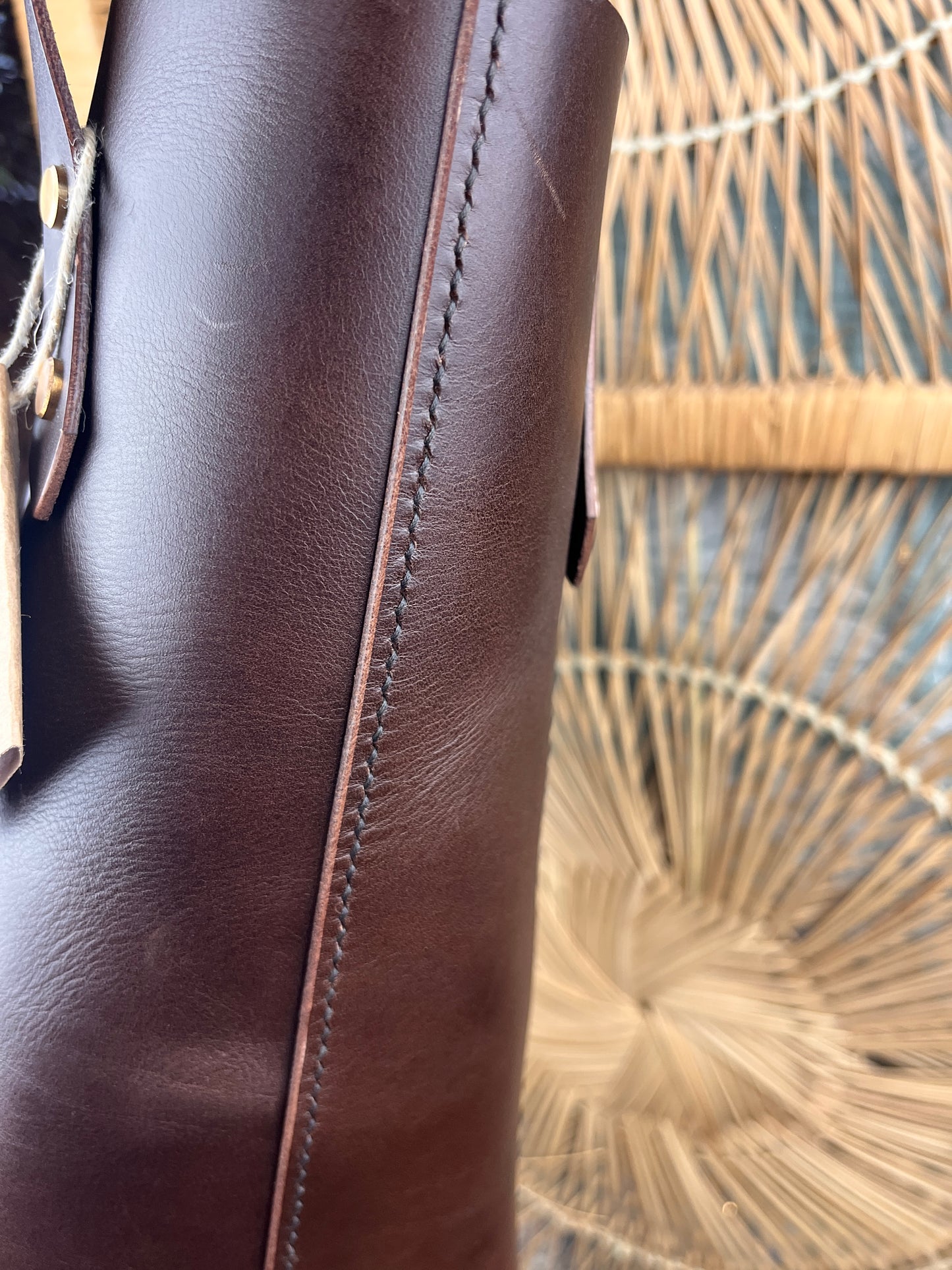 Close-up of a dark brown leather cylindrical case or handle with visible stitching, set against a woven straw or rattan background.