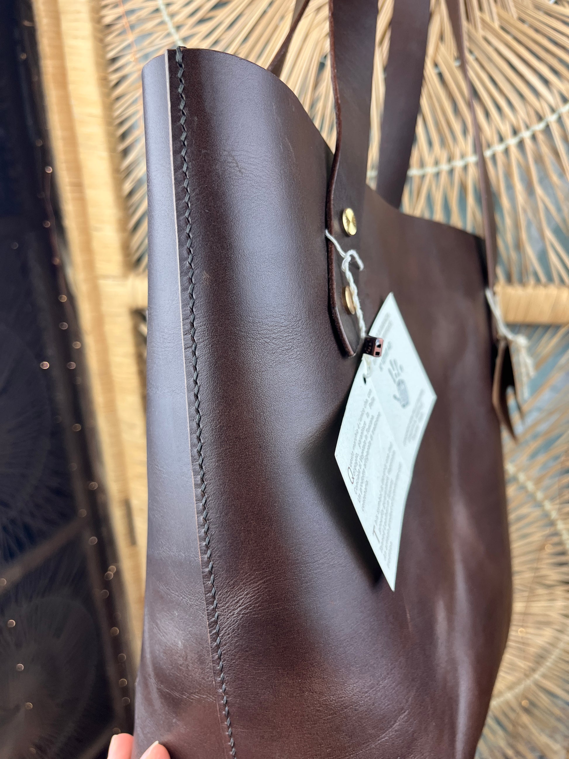 A close-up of a dark brown leather tote bag with a white product tag attached, held against a woven straw or rattan background.