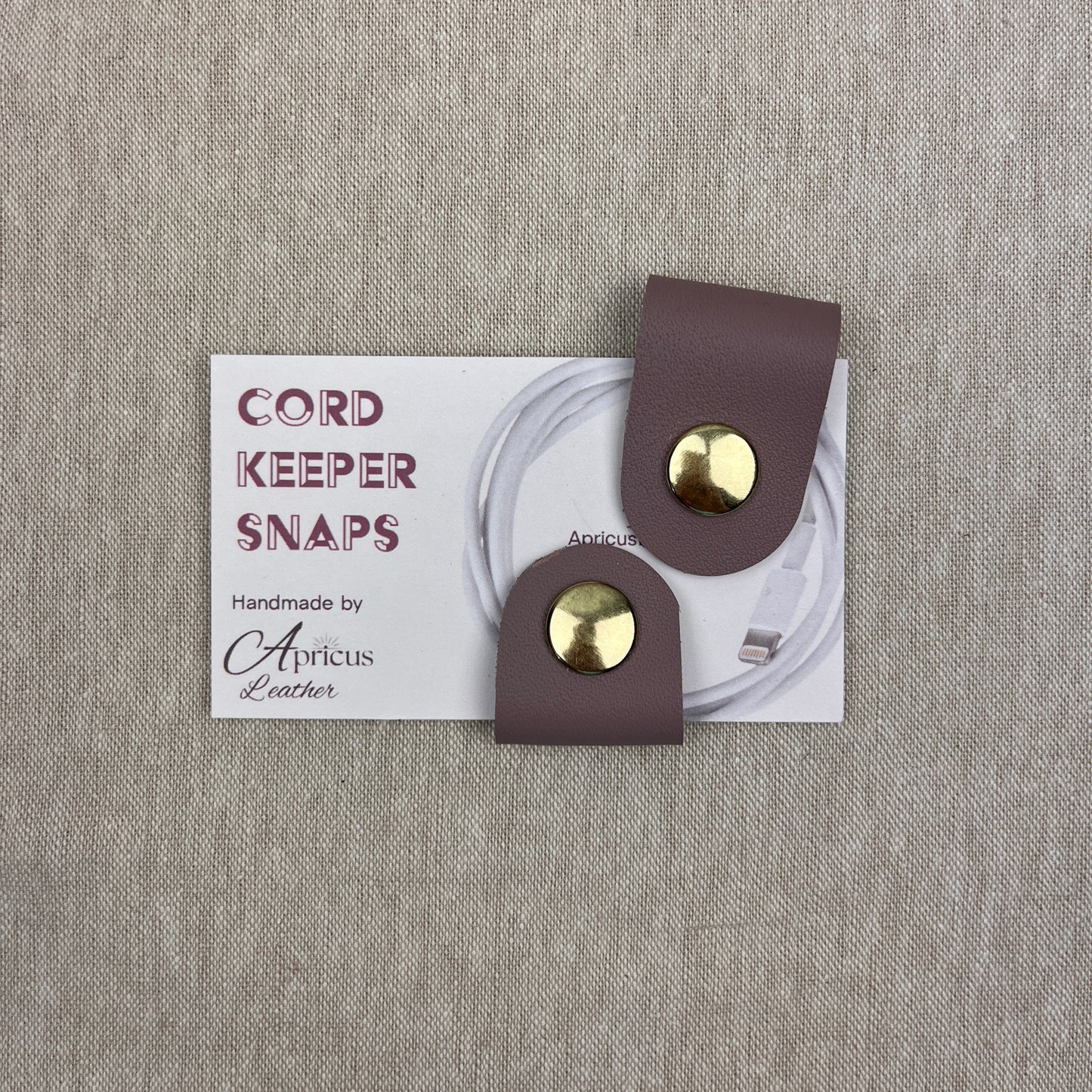 A pair of cord keeper snaps in a mauve leather finish with gold-tone metal snaps, shown on a white backing card that reads 'CORD KEEPER SNAPS' and 'Handmade by Apricus Leather', set against a textured beige linen background.