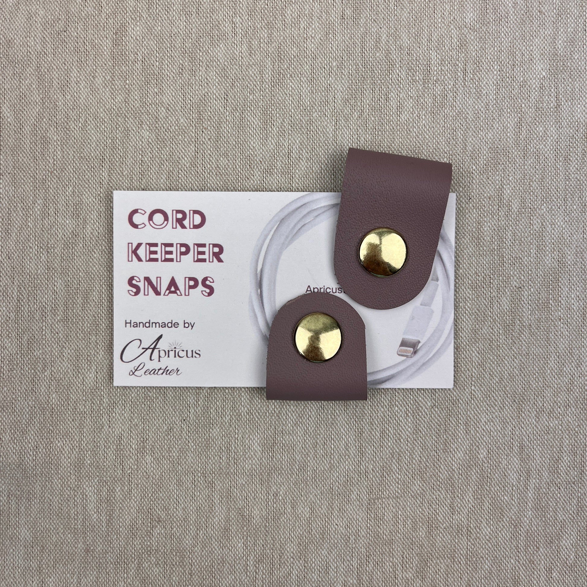 A pair of cord keeper snaps in a mauve leather finish with gold-tone metal snaps, shown on a white backing card that reads 'CORD KEEPER SNAPS' and 'Handmade by Apricus Leather', set against a textured beige linen background.
