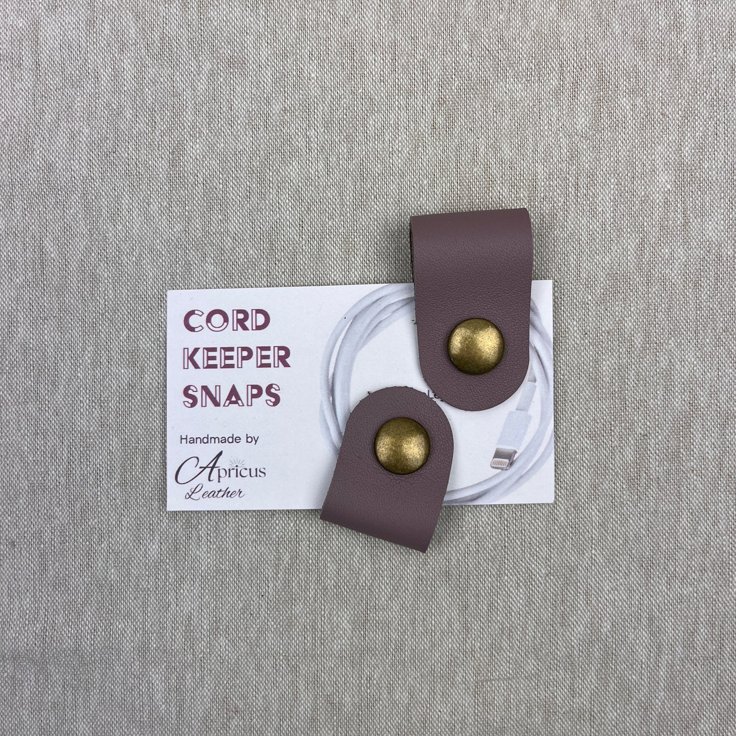 A pair of cord keepers with mauve-leather loops and gold-tone metal snaps, displayed on a white backing card over a textured gray linen background. The card reads 'CORD KEEPER SNAPS' and 'Handmade by Apricus Leather'.
