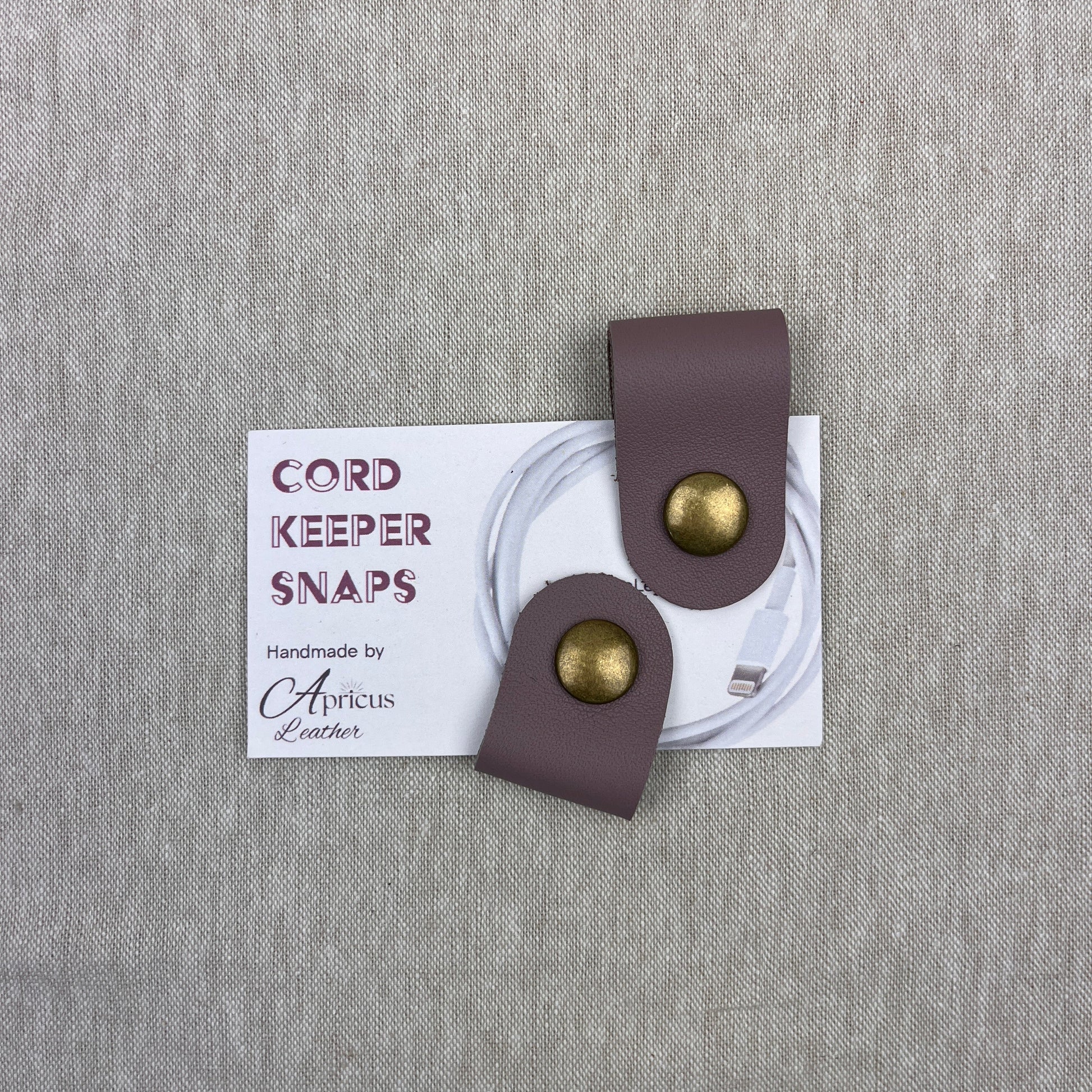A pair of cord keepers with mauve-leather loops and gold-tone metal snaps, displayed on a white backing card over a textured gray linen background. The card reads 'CORD KEEPER SNAPS' and 'Handmade by Apricus Leather'.