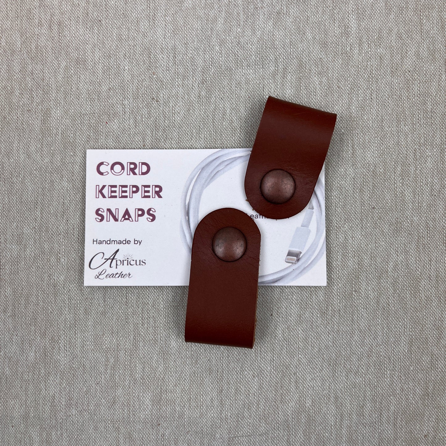 Two chestnut brown leather cord keepers with snap buttons, displayed on a white card labeled 'CORD KEEPER SNAPS' against a textured gray linen background.