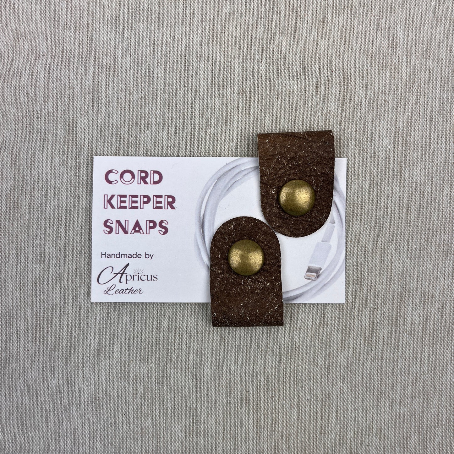 Two brown textured leather cord keepers with round gold-tone metal snaps, displayed on a white product card over a gray fabric background. The card reads 'CORD KEEPER SNAPS' and 'Handmade by Apricus Leather'.