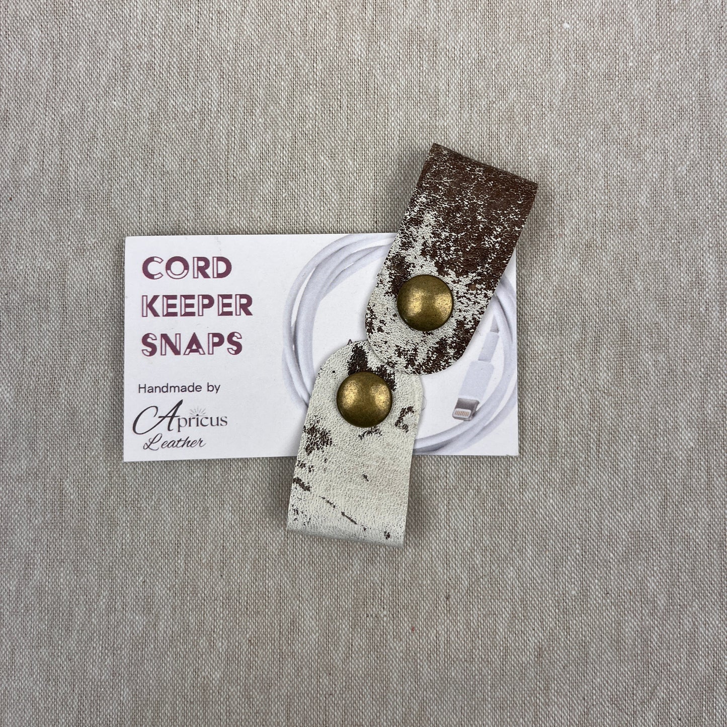 A cord keeper snap made of distressed leather in brown and off-white with brass snaps, displayed on its white product card against a textured gray linen background.