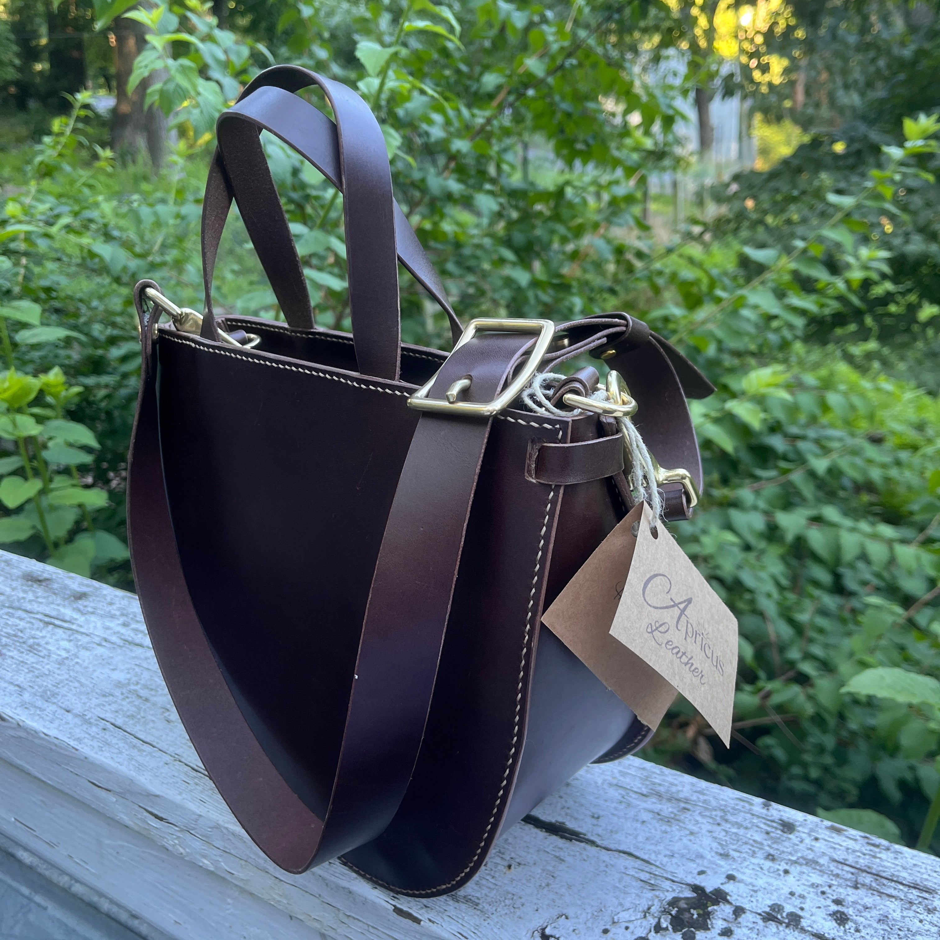 Brown leather handbag with a visible brand tag on a wooden surface with greenery in the background